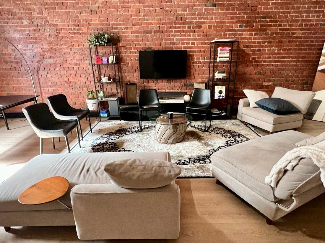 Beige modular lounges facing a wall-mounted TV, framed by a wall of exposed red brick